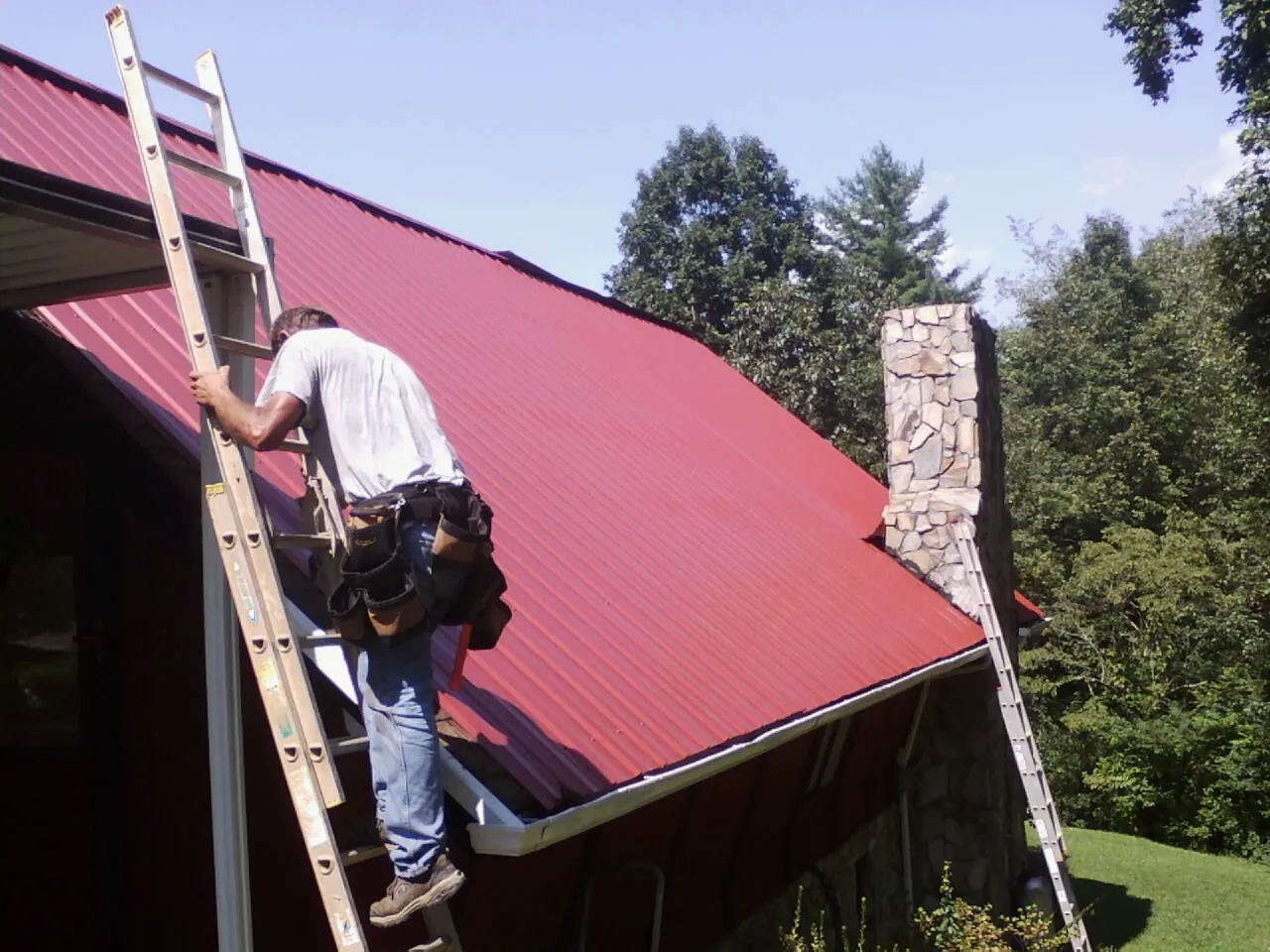 Roofer on a ladder beside a red metal roof and stone chimney — Soffit & Fascia Repair by Roofing Brothers in Kearney